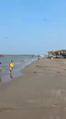 A woman in a yellow bikini runs along a crowded beach, passing under a flock of seagulls. The camera then focuses on another woman in a black bikini who is facing the camera and on the beach.
