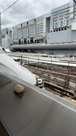 The video shows the front of a white Shinkansen train on tracks. A large, modern building with many windows is visible in the background. The train's windshield wipers are in the down position. The sky is overcast.