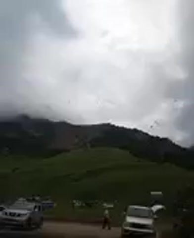 The video captures a landscape scene under a cloudy sky. In the foreground, several vehicles are parked on a dirt road. A silver SUV is on the left, and a white van is on the right. Further back, a few tents are set up, suggesting a campsite. A person is visible standing near the tents. The background features rolling green hills leading up to a mountain range partially obscured by clouds. Small dark specks, possibly birds, are scattered across the sky. The overall atmosphere is overcast and natural.