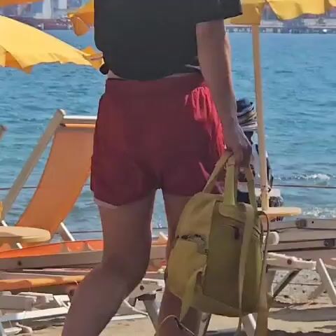 The video shows a beach scene with two orange lounge chairs and a yellow umbrella in the foreground. The ocean is visible in the background. A person, whose gender and race are not clearly discernible due to the low resolution and distance, is walking in the water behind the lounge chairs. The person is carrying a bag and appears to be wearing a hat. The person is walking towards the right side of the frame and out of view.
