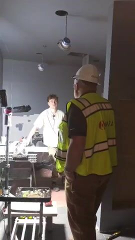 Two men are in a room under construction. One man, wearing a hard hat and a yellow safety vest with a logo, is facing away from the camera, looking at the other man. The second man, younger, wearing a white shirt and khaki pants, is facing the camera, gesturing with his hands. Both men appear to be engaged in a conversation. Tools and construction materials are visible in the foreground. The room has grey walls and a ceiling with exposed pipes and light fixtures.