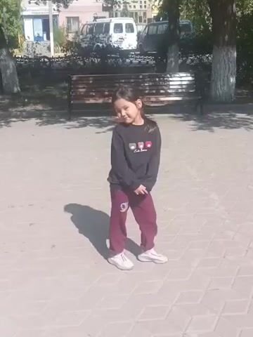 The video shows a young girl standing on a paved walkway. She is wearing a black long-sleeved shirt and maroon pants, with white shoes. The girl is looking down and to her right, with her head tilted. Behind her is a park bench, a tree, and some buildings and vehicles in the background. The sun is casting a shadow of the girl on the walkway.