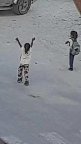 Two young children are standing on a dirt or sand-covered area. One child, wearing a white t-shirt and patterned pants, has their arms raised in the air. The other child, wearing a white t-shirt and jeans, has a backpack on and is walking away. A gray pickup truck is visible in the background. A woman in a black abaya and pink hijab is also present, facing the children. The lighting suggests it is daytime.