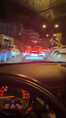The video shows the interior of a car at night, with the dashboard visible in the foreground. The car is in traffic, and the tail lights of a white Lamborghini are prominent in front. Other cars are also visible on the road, and buildings with lights are in the background. The scene appears to be in an urban environment.