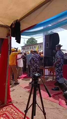 A group of people are gathered under a tent with red and white draped fabric and a blue and white striped fabric canopy. In the foreground, a black tripod with a camera is set up. Several individuals wearing yellow shirts and yellow hats are visible, some holding signs. There are also individuals in camouflage uniforms, possibly security personnel, present. One person in a brown robe is walking away from the camera. A large speaker is positioned to the right of the frame. The ground is covered with a red patterned rug in the foreground and dirt further back. In the background, there is a building with visible windows and a tower. The overall atmosphere appears to be an outdoor event or gathering.