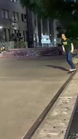 The video shows a young man skateboarding at an urban location in the evening or at night. He is wearing a dark green t-shirt, blue jeans, and light-colored sneakers. The video starts with him skating towards a metal rail, then jumping and grinding along it. After this, he continues skating on the paved surface. In the background, there are other people, buildings, street lamps, and some bicycles. The ground is concrete and there's graffiti on the walls. The overall impression is an urban, recreational scene with people enjoying skateboarding.