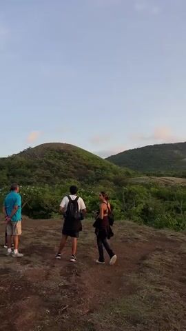 Two people stand on a grassy hill overlooking a valley covered in lush green trees. The sky above is a gradient of blue, with hints of clouds and the warm glow of the setting sun. The person on the left is wearing a white t-shirt, black shorts, and a backpack. The person on the right is wearing a dark tank top, dark pants, and also has a bag. The ground is a mix of dirt and dry grass. The camera pans slowly to the right, revealing more of the expansive, tree-filled landscape.