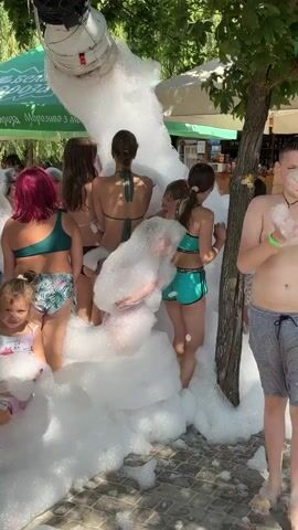 A group of children and adults are gathered outdoors, surrounded by trees and what appear to be tents or awnings in the background. A large machine suspended from a tree branch is dispensing a torrent of white foam, which is accumulating around the people. Many of the individuals are wearing swimwear. Some are actively playing in the foam, while others are observing the scene. The overall atmosphere appears to be festive and recreational, likely at an outdoor event or water park.