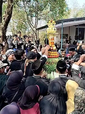 The video shows a crowd of people gathered outdoors around a tall, decorated structure, possibly a harvest offering or a ceremonial display. The structure is adorned with fruits and vegetables, topped with a golden, tiered ornament. Many individuals are dressed in black clothing and some wear traditional headwear, suggesting a cultural or religious event. Some people are reaching out towards the structure, possibly to take offerings or participate in a ritual. In the background, a building with a covered porch is visible, and trees are present, indicating an outdoor setting. A group of people in matching uniforms, some with musical instruments, are positioned on the left side of the frame. The overall atmosphere appears to be festive and communal.