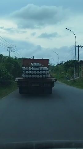 A flatbed truck is driving down a paved road under a cloudy sky. The truck is loaded with many cylindrical objects stacked horizontally. A motorcycle with two people on it is driving behind the truck, slightly to the right. Trees and utility poles line the sides of the road.