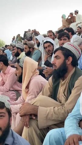The video shows a crowd of men and boys sitting and watching something off-camera. Most of the individuals are wearing traditional clothing, including turbans and caps. Some are wearing vests over their shirts. The setting appears to be outdoors, possibly a stadium or an open gathering area with tiered seating. The lighting suggests it is daytime. The individuals are looking in the same direction, indicating a shared focus of attention.