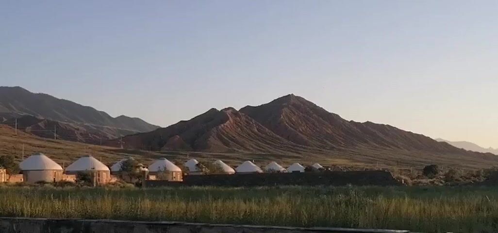 A wide shot shows a group of yurts arranged in a cluster in a grassy field. Beyond the yurts, a low wall runs horizontally across the frame, adorned with patterned decorations and what appears to be graffiti. In the background, rugged, tan-colored mountains rise against a clear, pale blue sky. The lighting suggests either early morning or late afternoon, with a soft glow illuminating the landscape. No people are visible in the frame.
