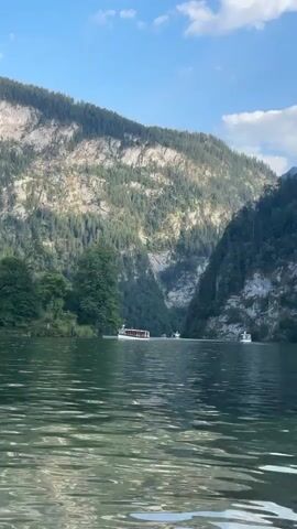 The video shows a wide, calm lake surrounded by steep, tree-covered mountains. The water is green and reflects the sky and mountains. There are several boats on the lake, including a larger white boat with red accents in the middle distance and a few smaller boats further away. The sky is blue with some scattered white clouds. The overall impression is one of a serene natural landscape.