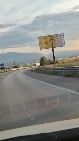 A dashboard view shows a highway in a rural area with mountains in the distance under a cloudy sky. A yellow billboard with the text "XO Park 16 km" and several logos is visible. The road curves to the right, and there are construction barriers with red and white markings along the side of the road. Sunlight streams through the clouds, creating visible rays.