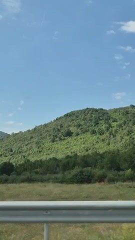 This is a vertical video of a scenic drive through a rural area in Dzhurovo, Bulgaria. The camera is positioned behind a metal guardrail, suggesting the view is from a vehicle. The foreground features the metallic guardrail, with hints of yellow wildflowers and green grass beyond it. The midground showcases a vast expanse of lush green hills, densely covered with trees. On the lower slopes of one of the hills, a few houses with tiled roofs are visible, indicating a small settlement. A tall wooden pole stands near one of the houses. The sky above is a clear, bright blue, dotted with sparse, wispy white clouds. The sunlight suggests it is daytime, likely mid-morning or afternoon, and the weather appears to be fair. The overall impression is one of tranquility and natural beauty. There is no visible text in the video.