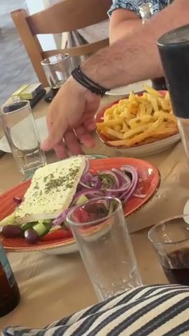 The video shows a table at a restaurant. On the table, there is a Greek salad with feta cheese, red onion, cucumber, olives, and tomatoes. There are also french fries, a bottle of Ouzo, and several glasses, some with drinks. The table is covered with a brown tablecloth. The lighting suggests it is daytime. The camera is mostly stationary, but there are some slight movements and blurs. The focus is primarily on the food and drinks.