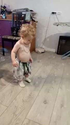 A child with curly blond hair and no shirt is playing with bubbles on a wooden floor. The child is wearing striped pants and white socks. In the background, there is a purple cabinet, a chair, and a television. The child is actively reaching for and interacting with the bubbles.