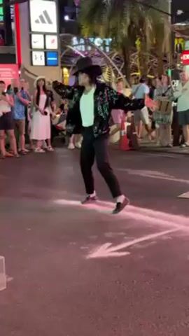 The video shows a Michael Jackson impersonator dancing in the street at night. The impersonator is wearing a black sequined jacket, black pants, a white t-shirt, and a black fedora. They are performing in front of a crowd of onlookers on a street marked by arrows of pink light. There are several shops and businesses visible in the background including an Exchange. A child is seen briefly dancing along with the performer.