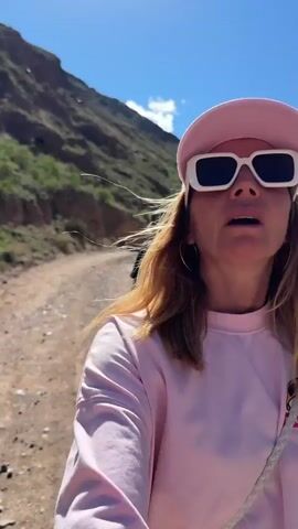 A woman wearing a pink shirt, pink hat, and large white sunglasses walks along a dirt road in a mountainous area. The sky is blue with a few clouds. There is a man walking ahead of the woman on the same road. The mountains are mostly brown and green.