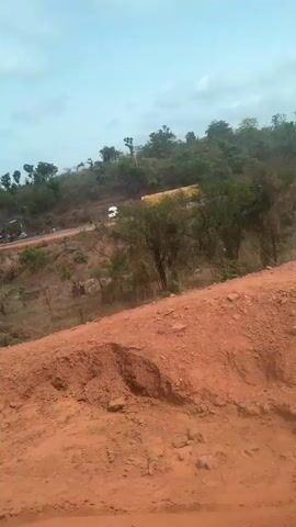From a medium distance, a construction site is visible from a dirt-covered hill. An excavator is parked to the left of the frame, working on the construction of a road. A white van and a large truck, possibly carrying construction materials, are on the road. The surrounding area is vegetated with small trees and dry grass. The sky is overcast. The camera is relatively still.