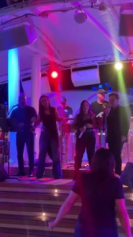A band performs on a stage under colorful lights. Five band members are on stage. One is singing into a microphone, and two others are singing or speaking into microphones. One band member plays a keyboard, and another plays drums. The band members wear dark clothing. Several people are in the audience, dancing. The stage has a staircase leading up to it. The environment appears to be outdoors at night.