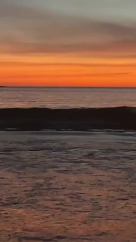 A video shows a sunset over the ocean. The sky is filled with vibrant orange and yellow hues, transitioning to a pale blue at the top. Gentle waves roll in towards a dark shore. The overall scene is serene and captures the beauty of a coastal sunset.