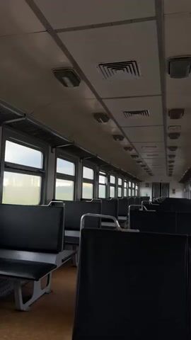 The video shows the interior of an empty train car. Rows of dark gray seats face each other across an aisle. The seats appear to be upholstered in vinyl or a similar material. Above the seats, there are luggage racks. The ceiling is a light gray color and features several light fixtures and air vents. Large windows line the side of the train car, offering a view of a blurry, overcast landscape with some green foliage. The lighting inside the train car is dim, creating a somber atmosphere. The train appears to be stationary.