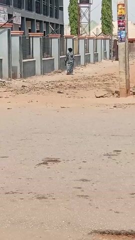 The video shows a daytime exterior shot of a street in what appears to be a developing country. The street is paved but marked with stains and discoloration. A dirt path runs adjacent to the street and behind a grey concrete fence which is next to a multi-story building.  An individual in a patterned dress walks along the dirt path away from the camera. A water tower stands in the background with two tall thin trees on either side. A utility pole with advertisements is to the right of the dirt path. A motorcycle is partially visible on the right side of the frame, parked near the utility pole. Another motorcycle drives past on the paved road.