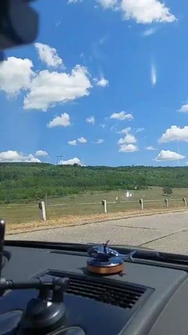 A dashboard-mounted smartphone and a small helicopter figurine are visible inside a car. The car is moving along a road with a grassy embankment and a wooded hill in the background. The sky is blue with scattered white clouds. Other vehicles, including a car and a van, can be seen on the road ahead. The camera is positioned to capture the view through the windshield.