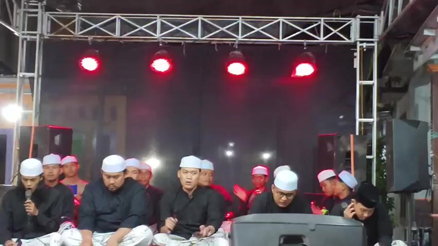 A group of men wearing white skullcaps and black shirts are sitting on the floor in front of a stage. They are holding microphones and appear to be singing or speaking. The stage has a black backdrop with stage lights above it. There are speakers on either side of the stage. Some of the men are holding phones. The overall setting appears to be an outdoor event at night.