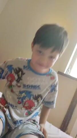 A young boy with short brown hair and a light complexion smiles at the camera. He is wearing pajamas featuring Sonic the Hedgehog characters. The background appears to be a bedroom with a window and a radiator visible. The lighting suggests it is daytime. The video is a close-up shot of the boy's face and upper body.