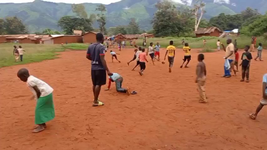 The video shows a group of children and adults playing soccer on a dirt field in what appears to be a rural village. Most of the children are dressed in casual clothes such as shorts and t-shirts, while some are shirtless. Two adults are present, observing and interacting with the children. One of the adults wears a light-colored shirt and jeans, while the other wears a dark t-shirt and shorts. Some of the children are actively playing soccer, running and kicking the ball, while others stand on the sidelines. One child is on the ground as the game continues. The background reveals a village with simple, possibly mud-brick buildings topped with thatched roofs. Lush green trees surround the village, and rolling mountains are visible in the distance under a cloudy sky.