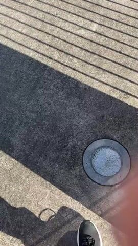 The video shows a first-person perspective of someone walking on a paved surface. Sunlight casts long shadows across the ground, indicating it is likely daytime. The person is wearing black pants and black athletic shoes with white soles. In the foreground, there are striped shadows from a fence or railing overhead. To the right of the frame, a circular, recessed light fixture is visible on the pavement, with some reflective liquid or debris inside it. The person's movement is casual, suggesting a walk rather than a brisk pace. The environment appears to be outdoors, possibly a park, walkway, or public space.