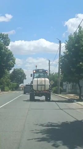 A blue tractor with a white tank is driving on a road. The tractor has a flashing yellow light on top and a license plate. There are trees and houses on the side of the road, and the sky is blue with some clouds. A car is visible behind the tractor. The road appears to be paved with asphalt and has white lane markings. There is a utility pole with wires on the right side of the road.