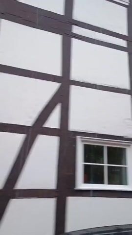 The video pans across the exterior of buildings. The first shot shows a white wall with dark brown timber framing and a small window. The second shot shows a stone wall in front of a row of houses with red tile roofs and a large evergreen tree. A dark-colored SUV is parked in front of one of the houses. The third shot shows a white building with multiple windows, some reflecting the sky and trees. The text "SOESTER ANZEIGE" is visible on the building. The fourth shot is a close-up of a white wall with several windows, some reflecting the sky and trees.