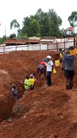 The video shows a group of people working at a construction site. The ground is red clay. There is an excavation in the foreground where some people are standing. Other people are carrying yellow containers of liquid up a slope. A man in a white hard hat and striped shirt seems to be supervising. Some buildings and trees are visible in the background under an overcast sky. There is a fence separating the construction site from the buildings. Some workers are wearing bright yellow safety vests. One person is wearing a red hard hat.