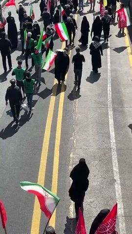 The media file depicts a procession of people walking down a paved road. The subjects are primarily individuals, many of whom are wearing black clothing, including headscarves and cloaks. Several individuals are carrying flags. The flags are predominantly red and green, with some featuring white. Some of the flags appear to have Arabic script on them, and some are identifiable as the flag of Iran due to its distinct red, white, and green horizontal stripes with a central emblem.

The setting appears to be an urban environment. The road is marked with yellow lines down the center, and there are sidewalks or elevated areas on either side. The sunlight suggests it is daytime, and the shadows indicate a bright, likely sunny, day. The activity is a march or procession, with the participants moving in the same direction. There is a sense of solemnity or purpose in the way they are walking.

Notable details include the diversity in the crowd, with some individuals wearing green vests, and others in full black religious attire. The flags are a significant element, suggesting a national or religious gathering. The presence of the Iranian flag strongly points to the location being Iran, specifically Tehran as indicated. The mood seems to be serious and respectful, with no overt signs of celebration or distress.

There is visible text on some of the red flags. While the script is in Arabic, it is difficult to read clearly in this media file. The overall scene is that of a public gathering in an urban setting, likely a demonstration or commemorative event, given the flags and the solemn atmosphere.