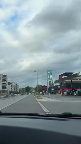 The video shows a street scene with a gas station prominently featured on the right. A tall, green digital sign displays gas prices: Super at 17.59, Super Plus at 18.89, and Diesel at 18.84. Cars are visible on the street and in the gas station area. To the left of the gas station, there's a shopping center sign that reads "copa" with a red circle and line through it, indicating "no entry." The sky is overcast with clouds. The camera appears to be inside a vehicle, looking forward through the windshield.