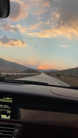 The video captures a first-person perspective from inside a car driving on a paved road through a mountainous landscape at sunset. The sky is filled with dramatic clouds, tinged with orange and pink hues, as the sun sets behind a mountain range in the distance, casting rays of light. The road ahead is empty, stretching towards the horizon. Debris, possibly insects or leaves, is visible on the windshield and in the air. The car's dashboard is partially visible in the foreground, showing some electronic displays. The overall mood is serene and contemplative, highlighting the beauty of the natural landscape during a sunset drive.