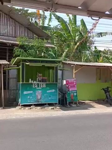 A roadside ice tea stand is visible on the left. To the right of the stand, a pink menu board is propped up. A man rides past on a red and white motorcycle, with another person behind him. A black motorcycle is parked to the right of the pink menu board. The area is lined with trees and buildings.