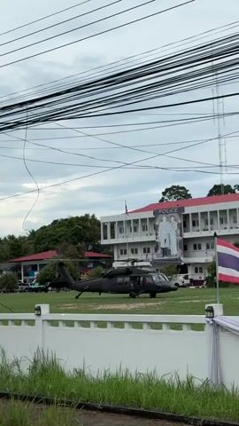 The video shows a black helicopter parked on the grass in front of a building with "POLICE" written on it. There is a Thai flag waving in the foreground. Power lines crisscross the sky above. A white wall and some green grass are in the immediate foreground. A poster of two people is attached to the front of the building. A telecommunications tower is visible behind the building. The sky is overcast.