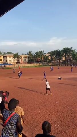 A group of soccer players wearing different uniforms are playing on a brown dirt field. Several players are standing, while one is on the ground, seemingly injured. One player in a white jersey with the number 10 on the back is gesturing with his arms. Buildings and palm trees can be seen in the background. There are spectators watching the game from the sidelines. The sky is a mix of blue and light clouds.