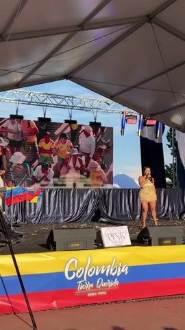 The video shows a performance on a stage outdoors. The stage is covered by a white tent structure. In the background, a large screen displays images of people in various colorful outfits, some playing musical instruments. In front of the screen, two performers are on stage. On the left, a male performer in a black outfit is dancing. On the right, a female performer in a gold, sequined dress is singing and dancing. The stage is decorated with a banner featuring the Colombian flag colors and the text "Colombia Tierra Querida Milan-Italia". Two large black speakers are visible on the stage. The lighting suggests it is daytime, with sunlight filtering through the tent. There are some confetti pieces scattered on the stage floor.