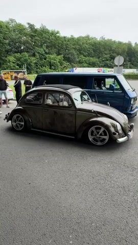 The video shows an outdoor event featuring several vintage Volkswagen vehicles. In the foreground, a customized brown Volkswagen Beetle with chrome wheels is parked. To its left, a light-colored Volkswagen Karmann Ghia with similar wheels is also visible. Behind the Beetle, a blue Volkswagen van and other vehicles can be seen. Several people are standing around the cars, observing them. Blue flags with white text are planted in the ground. The environment appears to be a parking lot or gathering area, with trees and a building in the background. The overall atmosphere seems to be a car show or a meet-up for Volkswagen enthusiasts.