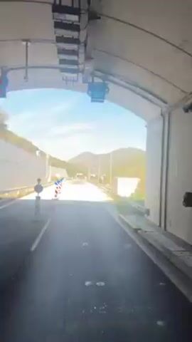The video shows a road winding through a mountainous area on a sunny day. The road has white concrete barriers on one side and a metal guardrail on the other. There are also striped red and white traffic barriers lining the side of the road. In the distance, mountains covered with trees can be seen under a bright blue sky with some scattered clouds. Streetlights are visible along the road. The road appears to be newly paved. The camera appears to be mounted on a vehicle driving forward.