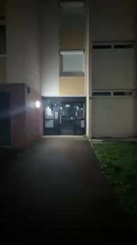 The scene is a nighttime exterior view of an apartment building entrance. The building is illuminated by dim exterior lights, casting shadows across the pathway leading to the entrance. The entrance consists of glass doors framed by dark metal. To the left of the entrance is a brick structure, and to the right is a patch of grass. The overall atmosphere is quiet and dark.