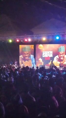 The video shows a crowd of people watching a performance on a stage. The stage features two large screens displaying an image of the performer, along with the text "SARKIIN DARLA".  The performer is dressed in a light blue outfit. There are bright spotlights shining on the stage. The audience is diverse. The scene appears to be an entertainment event, possibly a comedy show or concert.