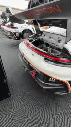 Here is a description of the video:

The video shows a white Porsche GT4 Clubsport race car with its hood open, revealing the engine compartment. The car is parked on a grey and white geometric-patterned floor, likely in a garage or pit area. The car has racing livery, including stripes and sponsor logos. The back of the car has a large rear wing with "EVOLUTION" written on it.

There's another similar race car parked behind it. A black tool cabinet with "GP" on the side is standing next to the car. Other cars and people can be seen in the background, suggesting a race event or car show. The camera is slowly panning leftward.