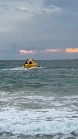A yellow inflatable tube, likely a banana boat, is being pulled across the ocean by a jet ski. There are people on the inflatable and on the jet ski. The water is choppy with waves breaking on a shore in the foreground. The sky is overcast with hints of pink and orange from a setting sun.