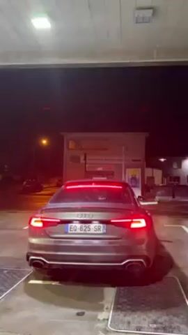 The video shows the rear of a gray Audi RS5 parked under a gas station canopy at night. The car's taillights and a light bar above the trunk are illuminated. The license plate number is EQ-625-SR. In the background, there is a building with "Laboratoire de Biologie Medicale" printed on it. The car's engine revs, producing an audible sound. The ground is wet, and there are grid-like covers on either side of the vehicle.